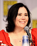 A woman with black hair, tied back, smiling, and sitting behind a microphone.