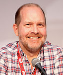 A man with closely shaven hair, and slight stubble, looking to the side slightly with his eyes, behind a microphone.