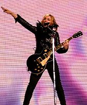 A woman in black clothing holding a guitar and standing behind a microphone stand with one arm extended straight into the air. In the background is a screen with shades of pink and purple.
