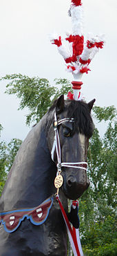 Tête d'un grand cheval de bois de couleur brune foncée, surmontée d'un plumeau rouge et blanc.