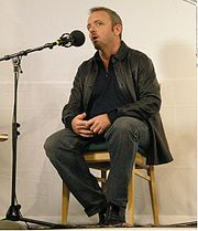 Photographie de Dennis Lehane, assis, parlant dans un micro, lors de la présentation de l’un de ses films en 2006