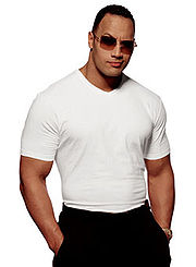 Vue de face du catcheur The Rock. Il est vêtu d'un T-shirt blanc et d'un pantalon noir.