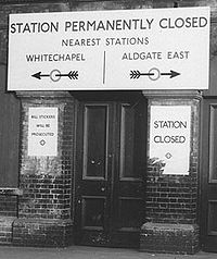 Une étroite porte en bois entre deux contreforts en briques. Un grand panneau au-dessus « Station fermée de façon permanente. Station la plus proche, Whitechapel / Aldgate East ». Les flèches indiquent les directions de ces deux stations.