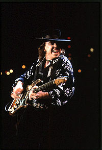 Photographie de Stevie Ray Vaughan en concert, de profil, tête tournée vers l'objectif, en train de jour de la guitare.