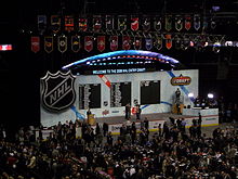 Photo couleur d'une scène avec le logo de la LNH, surmontée des fanions des équipes de la LNH et devant de nombreux spectateurs.