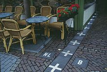 Ligne de pavés gris surmontés de croix blanches, qui délimite la frontière au pied d'un café.