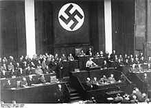 Photographie en noir et blanc prise lors du discours d’Adolf Hitler devant les membres du Reichstag le 23 mars 1933. La moitié supérieure de l’image est dominée par une immense bannière verticale à l’emblème de la croix gammée. Dans la moitié inférieure de l’image, à gauche et à droite, trois rangées de bancs dont les occupants ne sont pas visuellement identifiables. Au centre, de bas en haut, le bureau des sténographes, la tribune, et le siège du président du Reichstag, occupé par Hermann Göring. A la tribune, Adolf Hitler est revêtu de l’uniforme brun des SA ; on distingue à peine le baudrier qui lui barre la poitrine et le brassard à croix gammée à sa manche gauche. Hitler est debout, très légèrement penché vers l’avant ; il a les bras écartés et saisit des deux mains les bords du pupitre.