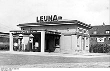 Photographie en noir et blanc montrant une station-service. Le mot « LEUNA » apparaît au-dessus de l'immeuble, alors que les expressions « Deutches Benzin » et « Tankdienst » décorent certaines parties de l'immeuble.