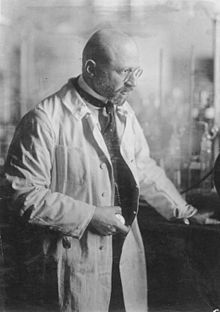 Photographie d'un homme se tenant debout dans un laboratoire. Sa main droite tient une montre de poche. L'homme porte une blouse, est chauve, porte des lunettes ainsi qu'une barbe.