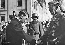 Photographie en noir et blanc prise à Potsdam le 21 mars 1933. À gauche de l'image, Adolf Hitler, vêtu d'une redingote, serre la main du président von Hindenburg, en s'inclinant vers lui. À droite, Hidenburg domine Hitler d'une bonne tête et se tient droit. Ganté, il porte le grand uniforme de maréchal prussien, arborant de nombreuses décorations dont la croix de fer, et un casque à pointe. Au centre et à l'arrière plan, entre les deux hommes, un militaire avec le lourd casque de combat de l'armée allemande