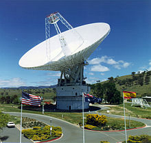 Une antenne parabolique blanche pointée vers le ciel avec les drapeaux américain, australien et espagnol au premier plan