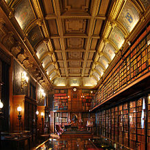 Vue des deux murs de la bibliothèque parcourus par une passerelle à droite et en face, de vitrines d'expositions au centre et d'armoires de livres à gauche. Le tout est surplombé d'un plafond à caissons