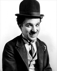 Un homme souriant portant une petite moustache, un chapeau-melon, une cravate et un manteau