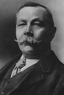 La photographie noir et blanc montre un homme d'une quarantaine d'années avec une moustache en guidon. Il porte un costume et une cravate.
