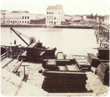 Une vieille photo montrant des tas de matériaux de construction sur la berge d'un fleuve avec de grands bâtiments blancs qui bordent la rive opposée