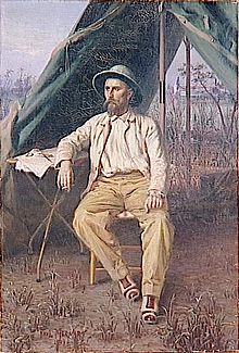 Un homme barbu en tenue coloniale, assis devant une tente de toile, le bras reposant sur une table à trépied métallique