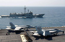 Deux chasseurs F/A-18 sur le pont d'envol, prêts au catapultage. Le HMAS Newcastle australien est visible en arrière-plan.