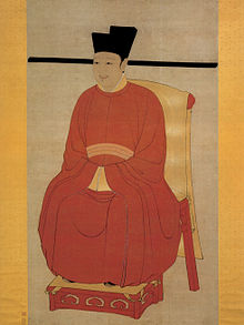 Peinture d'un homme assis sur une chaise rouge recouverte d'un drap jaune. L'homme porte un chapeau noir, une petite moustache et une barbiche. Il est vêtu d'une large robe rouge et d'une ceinture. Il croise ses mains, sous ses larges manches, au niveau de son bas ventre.