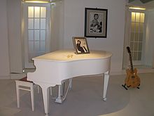 Un piano blanc dans une pièce blanche, entouré d'une des guitares emblématiques de Lennon, de sa paire de lunettes rondes et de deux photos de lui.