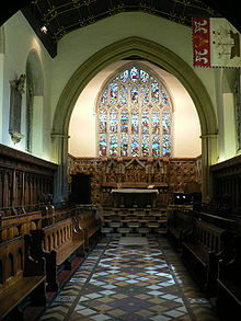 Photo de la chapelle ; La même chapelle avec l'arche du chœur plus large  et un retable de pierre en dessous d'un grand vitrail. Un pavillon décoré d'un navire est accroché en haut de la paroi de droite