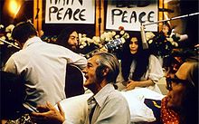 John Lennon (avec une guitare) et Yoko Ono, dans un lit entouré de gens dans une chambre d'hôtel.