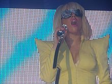 Une femme vêtue d'un veston jaune est visibile. Ses cheveux sont en forme de bob et elle porte une paire de lunettes. Elle tient un microphone dans sa main droite qui va jusqu'à sa bouche. Le fond de l'image montre un écran avec des motifs indigos et blancs.