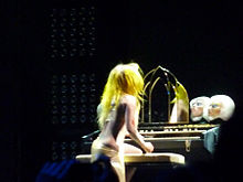 Gaga portant une perruque jaune et une tunique de couleur chaire positionnée devant un orgue qui est agrémenté de diverses têtes de mannequin.