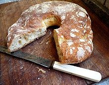 La photographie représente une couronne de pain. Le quart enlevé montre une mie aux bulles irrégulières sous une croûte dorée et farinée. Le pain repose sur une planche à pain en bois avec un couteau à dent à manche de plastique blanc.