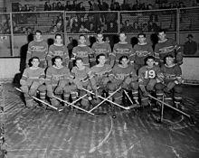 Photo noir et blanc des 14 joueurs des Canadiens de Montréal qui posent sur deux rangs sur la patinoire devant un grillage qui les sépare des gradins et des spectateurs.