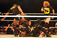 Sur un ring, trois membres du Nexus — Justin Gabriel (de dos), Wade Barrett (masqué) et Heath Slater — attaquent le catcheur Goldust, à terre.