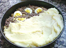 Plat rond contenant un pastel de papa en cours de préparation dans lequel on voit la couche inférieure de viande hachée de couleur brune, avec quelques quartiers d'œuf dur (jaune et blanc), aux deux tiers recouverte d'une couche de purée de couleur blanche.