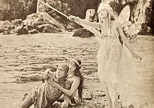À la gauche de la photo, un chevalier qui semble blessé est supporté par une femme, les deux étant assis par terre. À la droite, une fée en position debout brandit une baguette.
