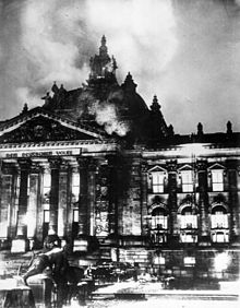 Photographie en noir et blanc du Reichstag en flammes, dans la nuit du 27 au 28 février 1933. L'image est de mauvaise qualité et ne permet pas de distinguer les détails. À la gauche de l'image, le fronton du palais du Reichstag est immédiatement identifiable et l'inscription qui y est gravée (Dem deutschen Volke-Au peuple allemand) tout à fait lisible ; de la coupole qui le surmonte, s'échappent des volutes de fumée. À la droite de l'image, une aile du palais, dont toutes les fenêtres laissent apparaître des flammes qui témoignent de la violence de l'incendie