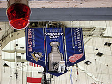 Photo de la bannière de la Coupe Stanley pendue au plafond de la patinoire représentant la coupe Stanley encadrée des logos des Penguins et des Red Wings associés au nom de leur conférence respective.