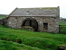 La photo montre un bâtiment en pierre d'un âge certain. Au milieu du bâtiment se trouvent les restes d'une roue en bois.