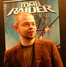 Un homme aux cheveux courts et avec des lunettes se tient près d'un panneau publicitaire. Il porte un t-shirt et une veste marrons.