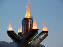 Quatre piliers avec la flamme à leurs sommets entourant un cinquième piller, seul, au milieu, avec aussi la flamme au sommet. Le fond est le ciel avec la montagne.