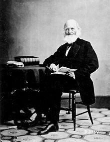 La photographie noir et blanc montre un homme âgé dans un costume noir. Il porte une barbe blanche imposante, et tient un livre ouvert, assis sur une chaise. Il est accoudé à une table sur laquelle repose un épais livre noir et une liasse de papier.