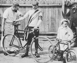 A black and white photograph of a man holding his bicycle and a little boy with a little bicycle, being looked upon by two other men.