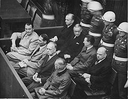 La photographie en noir et blanc de 1946 montre les accusés dans un box du tribunal ; derrière eux, quatre soldats casqués de blanc et en uniforme d'apparat les surveillent.
