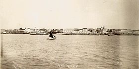 Photographie d'un plan d'eau avec un petit voilier au premier plan et des bâtiments le long du rivage en arrière-plan