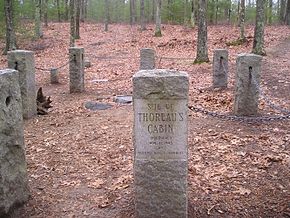 Lieu où Thoreau édifia sa hutte et délimité par des stèles en pierre. Le sol est recouvert de feuilles d'automne. Au fond, à l'arrière-plan : les bois de Walden. Une stèle affichant « Site of Thoreau's cabin » est au premier plan.