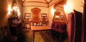 Petit salon dans lequel se trouve une table au centre, une chaise et une cheminée sur la gauche, une porte entourée de deux meubles au fond et, sur la droite, un sofa surmonté d'un miroir ainsi qu'un paravent, le tout dans le style art nouveau
