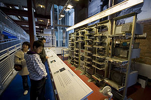 Une série de 7 grands casiers de métal remplis d'appareils électroniques devant un mur de briques. Un panneau au-dessus de chaque casier décrit le rôle des composants qu'il contient. À gauche de l'image, trois visiteurs lisent les informations.