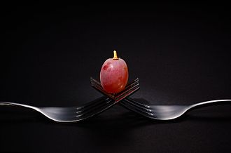 La photographie couleur montre deux fourchettes aux dents entremêlées. Un grain de raisin rosé est délicatement posé sur les dents. Le fond gris sombre fait ressortir l'éclat métallique brillant des fourchettes. Le grain de raisin est représenté comme un joyau dans son écrin.